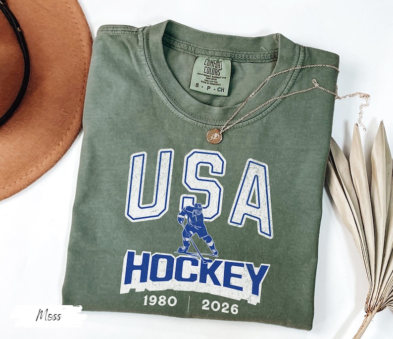 May include: An olive green t-shirt with the words "USA HOCKEY" in white and blue lettering. A hockey player graphic is featured below the word "USA". The shirt also has the years "1980" and "2026" printed on it. A gold necklace is draped across the shirt.
