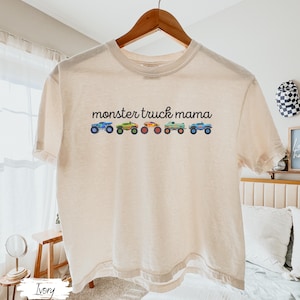 May include: Ivory-colored t-shirt with the text "monster truck mama" above a row of colorful monster truck illustrations. The trucks are blue, green, orange, and teal. The shirt is hanging on a wooden hanger.