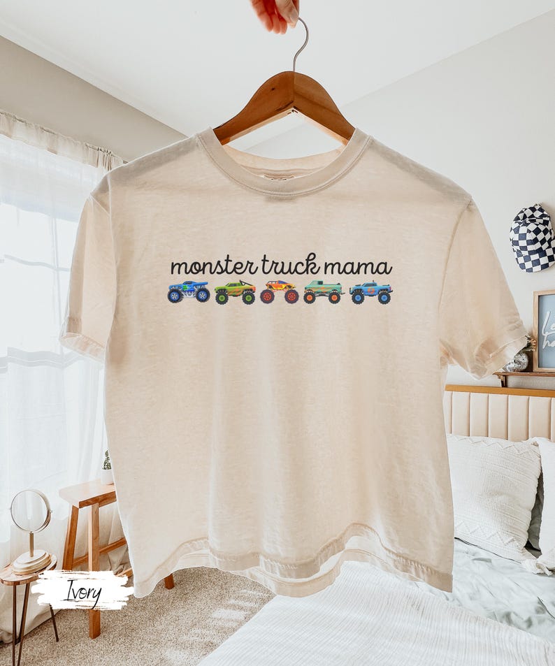 May include: Ivory-colored t-shirt with the text "monster truck mama" above a row of colorful monster truck illustrations. The trucks are blue, green, orange, and teal. The shirt is hanging on a wooden hanger.