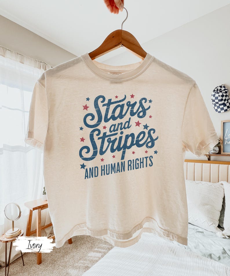 Puede incluir: Camiseta de color marfil con el texto "Stars and Stripes AND HUMAN RIGHTS" en azul y rojo. El dise&ntilde;o incluye estrellas. La camiseta est&aacute; colgada en una percha de madera.