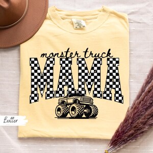 May include: Butter-colored t-shirt with the text "monster truck MAMA" in black. The word "MAMA" is in a checkered pattern, and a monster truck graphic is below the text. A brown hat is in the upper left corner.