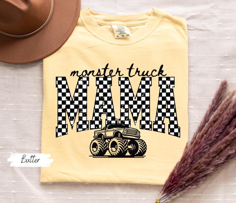 May include: Butter-colored t-shirt with the text "monster truck MAMA" in black. The word "MAMA" is in a checkered pattern, and a monster truck graphic is below the text. A brown hat is in the upper left corner.
