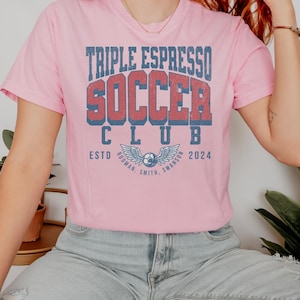 Op de afbeelding: Een roze T-shirt met de tekst "Triple Espresso Soccer Club" in een vintage lettertype. De tekst is in rood en blauw met een voetbal en vleugels eronder. De tekst "Est. 2024" staat onder de voetbal en vleugels.