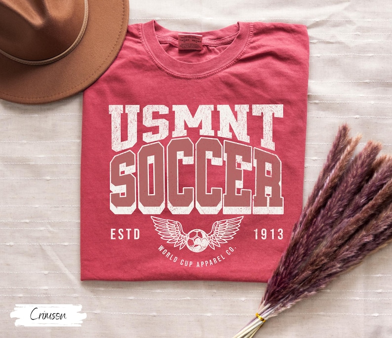 Może przedstawiać: Karmazynowa koszulka z napisem "USMNT SOCCER" w postarzanym białym i czerwonym kroju pisma. Poniżej grafika piłki nożnej ze skrzydłami oraz słowa "ESTD 1913" i "WORLD CUP APPAREL CO."
