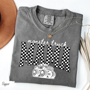 May include: A gray Comfort Colors t-shirt with the words "monster truck MAMA" in white. The word "MAMA" is in a checkered pattern. A white monster truck graphic is below the text. A gold necklace is on the shirt.