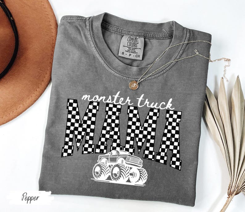 May include: A gray Comfort Colors t-shirt with the words "monster truck MAMA" in white. The word "MAMA" is in a checkered pattern. A white monster truck graphic is below the text. A gold necklace is on the shirt.