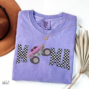 May include: Violet Comfort Colors t-shirt with a graphic of a pink monster truck and the word "MAMA" in a checkered pattern. A gold necklace and a brown hat are also visible.