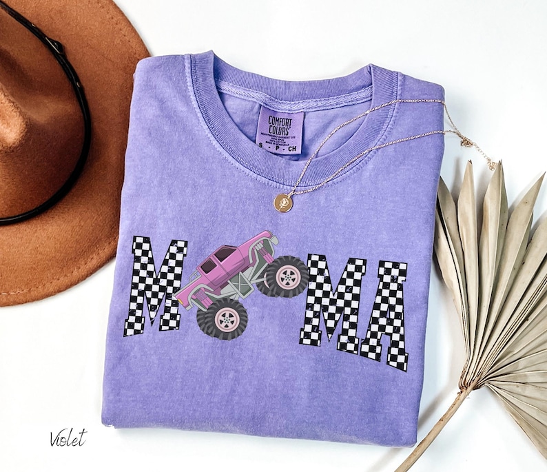 May include: Violet Comfort Colors t-shirt with a graphic of a pink monster truck and the word "MAMA" in a checkered pattern. A gold necklace and a brown hat are also visible.