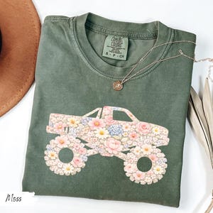 Op de afbeelding: Mosgroen t-shirt met een bloemen monster truck ontwerp. De truck bestaat uit roze, witte en blauwe bloemen. Het shirt heeft een ronde hals en korte mouwen. Een gouden ketting met een hanger is ook zichtbaar.