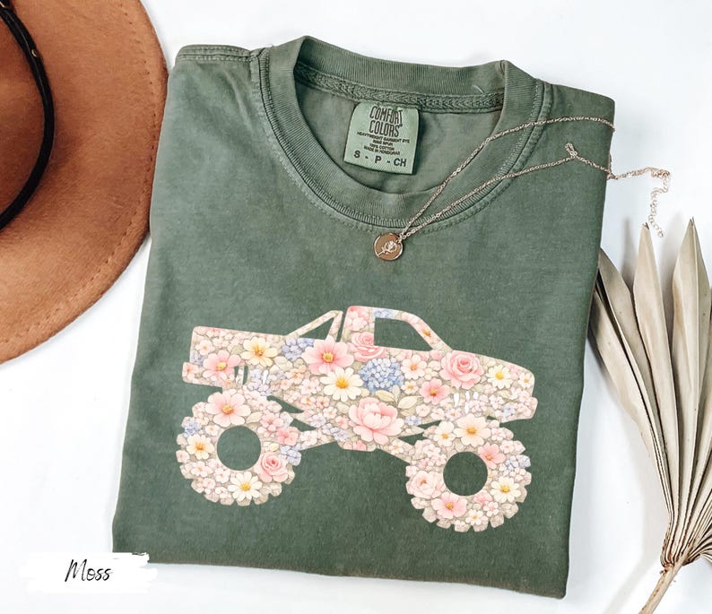 Op de afbeelding: Mosgroen t-shirt met een bloemen monster truck ontwerp. De truck bestaat uit roze, witte en blauwe bloemen. Het shirt heeft een ronde hals en korte mouwen. Een gouden ketting met een hanger is ook zichtbaar.