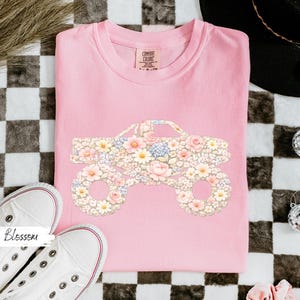 Op de afbeelding: Een lichtroze Comfort Colors sweatshirt met een bloemen monster truck ontwerp. De truck is gevuld met roze, witte en gele bloemen. Witte sneakers met rode rand zijn op de voorgrond.