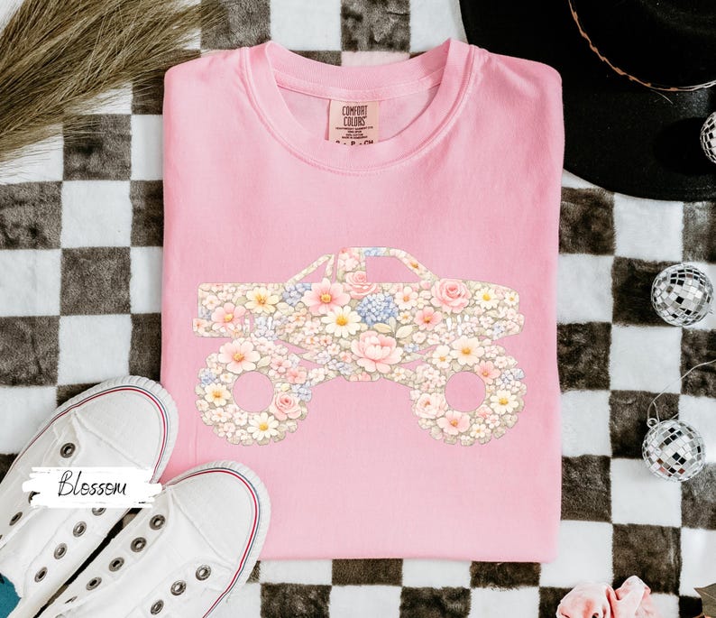 Op de afbeelding: Een lichtroze Comfort Colors sweatshirt met een bloemen monster truck ontwerp. De truck is gevuld met roze, witte en gele bloemen. Witte sneakers met rode rand zijn op de voorgrond.
