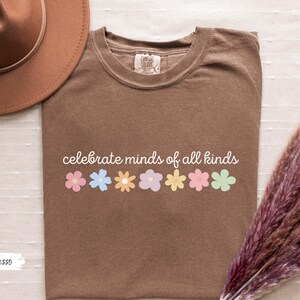 May include: A brown t-shirt with the text "celebrate minds of all kinds" printed in white. A row of colorful flowers are printed below the text.
