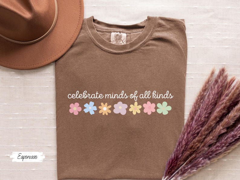 May include: A brown t-shirt with the text "celebrate minds of all kinds" printed in white. A row of colorful flowers are printed below the text.