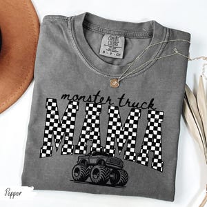Op de afbeelding: Een grijs t-shirt met de tekst "monster truck MAMA" in zwart-witte geblokte letters. Een zwarte monster truck afbeelding staat onder de tekst. Het shirt heeft een ronde hals en korte mouwen.