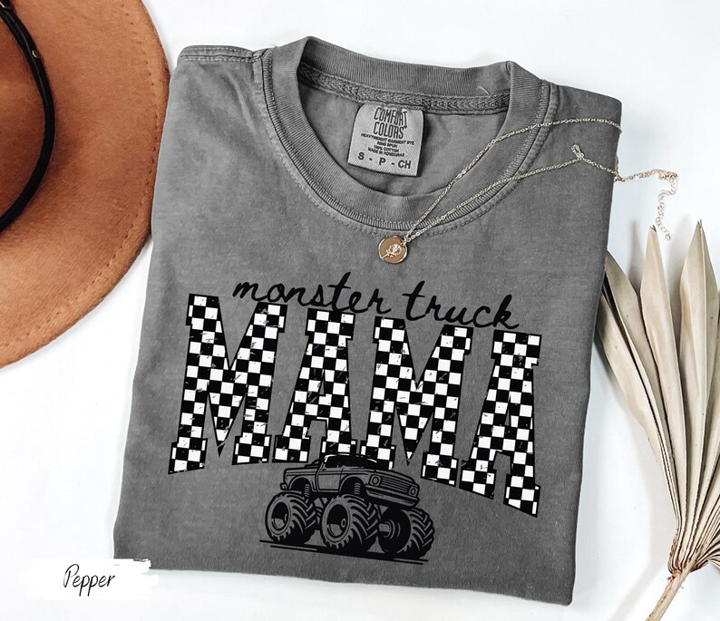 Op de afbeelding: Een grijs t-shirt met de tekst "monster truck MAMA" in zwart-witte geblokte letters. Een zwarte monster truck afbeelding staat onder de tekst. Het shirt heeft een ronde hals en korte mouwen.