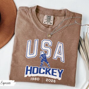 May include: Espresso-colored t-shirt with "USA HOCKEY" in white and blue lettering. Features a hockey player graphic and the years 1980 and 2026. A silver necklace with a pendant is draped across the shirt.