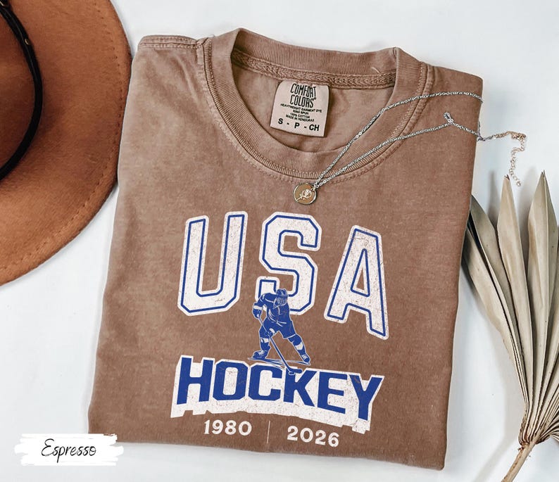 May include: Espresso-colored t-shirt with "USA HOCKEY" in white and blue lettering. Features a hockey player graphic and the years 1980 and 2026. A silver necklace with a pendant is draped across the shirt.