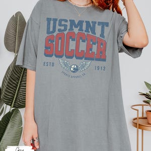 May include: A grey t-shirt featuring the text "USMNT SOCCER" in red and blue, with a soccer ball and wings graphic. The shirt also has "ESTD 1913" and "SPORTS APPAREL CO." printed on it. The word "Grey" is written at the bottom.