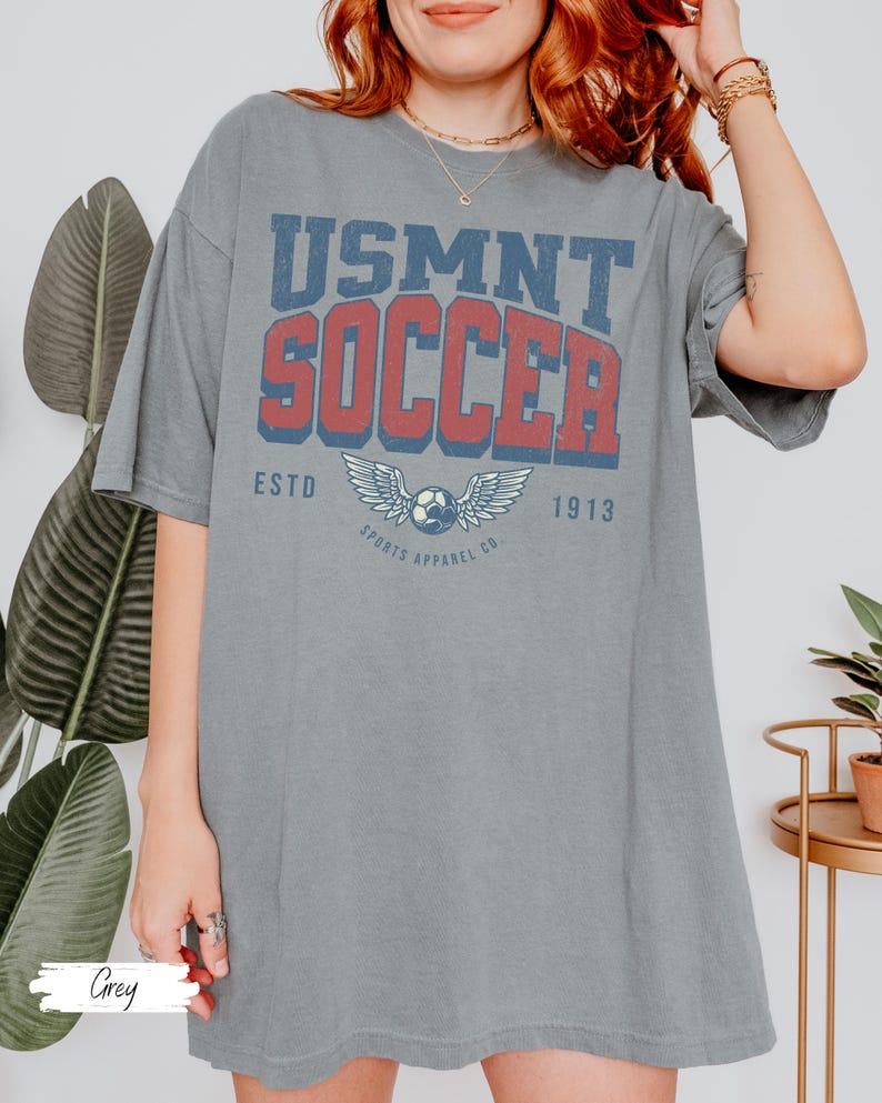 May include: A grey t-shirt featuring the text "USMNT SOCCER" in red and blue, with a soccer ball and wings graphic. The shirt also has "ESTD 1913" and "SPORTS APPAREL CO." printed on it. The word "Grey" is written at the bottom.