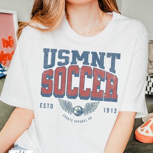 May include: White t-shirt featuring a distressed USMNT SOCCER graphic in blue and red. The design includes a soccer ball with wings and the text "ESTD 1913 SPORTS APPAREL CO."