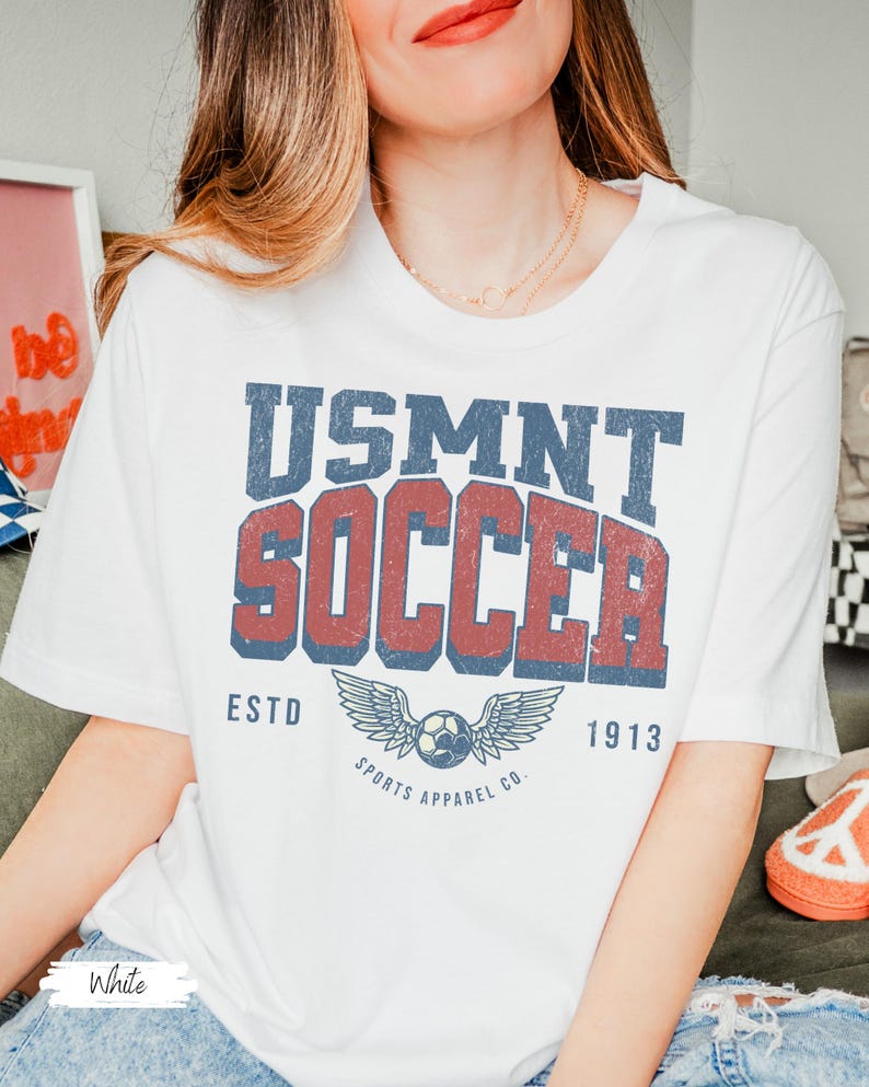 May include: White t-shirt featuring a distressed USMNT SOCCER graphic in blue and red. The design includes a soccer ball with wings and the text "ESTD 1913 SPORTS APPAREL CO."