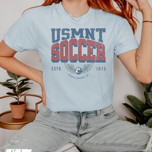 May include: Pale blue t-shirt featuring the text "USMNT SOCCER" in red and blue, with a soccer ball and wings graphic. The shirt also has the text "ESTD 1913" and "SPORTS APPAREL CO."