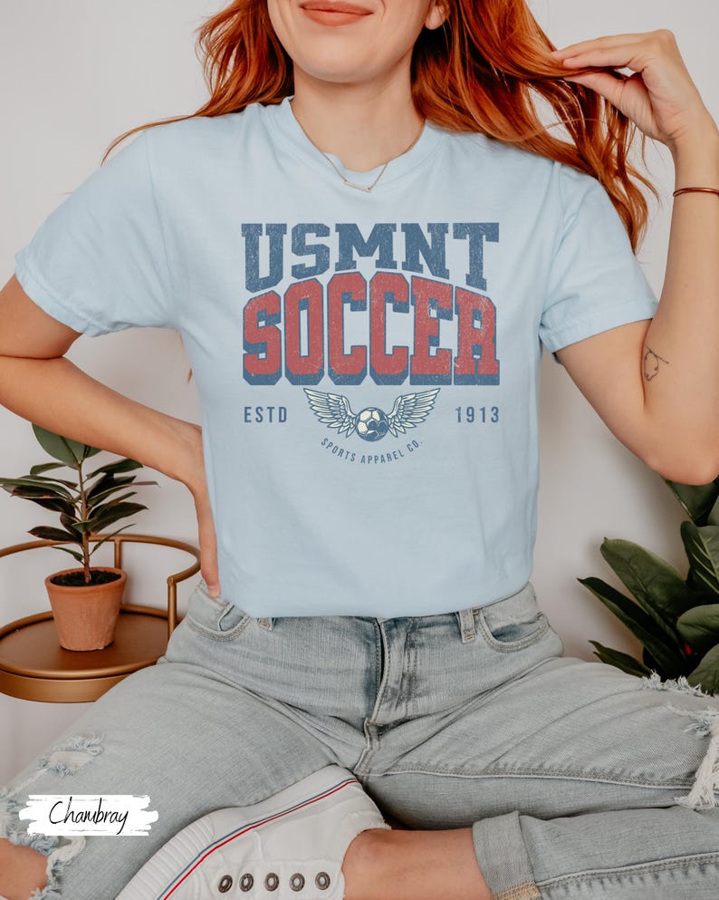 May include: Pale blue t-shirt featuring the text "USMNT SOCCER" in red and blue, with a soccer ball and wings graphic. The shirt also has the text "ESTD 1913" and "SPORTS APPAREL CO."