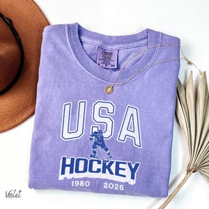 May include: A violet Comfort Colors t-shirt with a white and blue graphic that says "USA HOCKEY" with a hockey player illustration. The shirt also has the years "1980" and "2026" printed on it. A gold necklace is draped across the shirt.