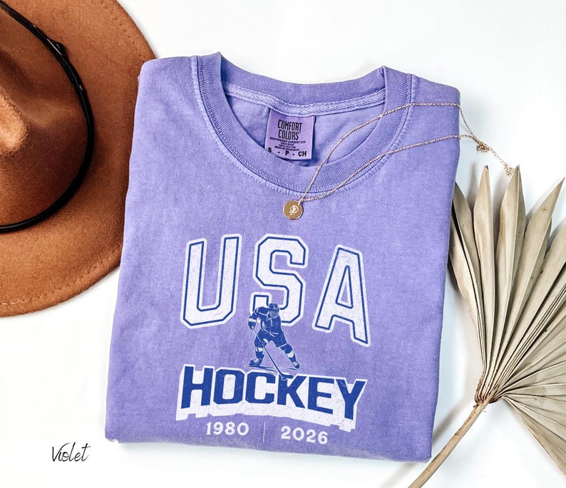 May include: A violet Comfort Colors t-shirt with a white and blue graphic that says "USA HOCKEY" with a hockey player illustration. The shirt also has the years "1980" and "2026" printed on it. A gold necklace is draped across the shirt.