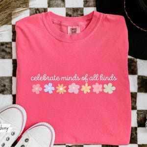 May include: A pink t-shirt with the text "celebrate minds of all kinds" and a row of colorful flowers printed on the front.