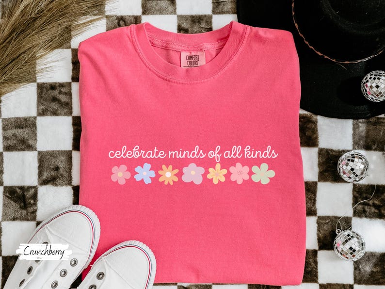 May include: A pink t-shirt with the text "celebrate minds of all kinds" and a row of colorful flowers printed on the front.