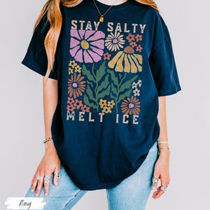 Op de afbeelding: Marineblauwe t-shirt met een bloemenontwerp en de tekst "STAY SALTY MELT ICE". De afbeelding toont roze, gele, oranje en witte bloemen met groene bladeren. Het shirt heeft een relaxte pasvorm.