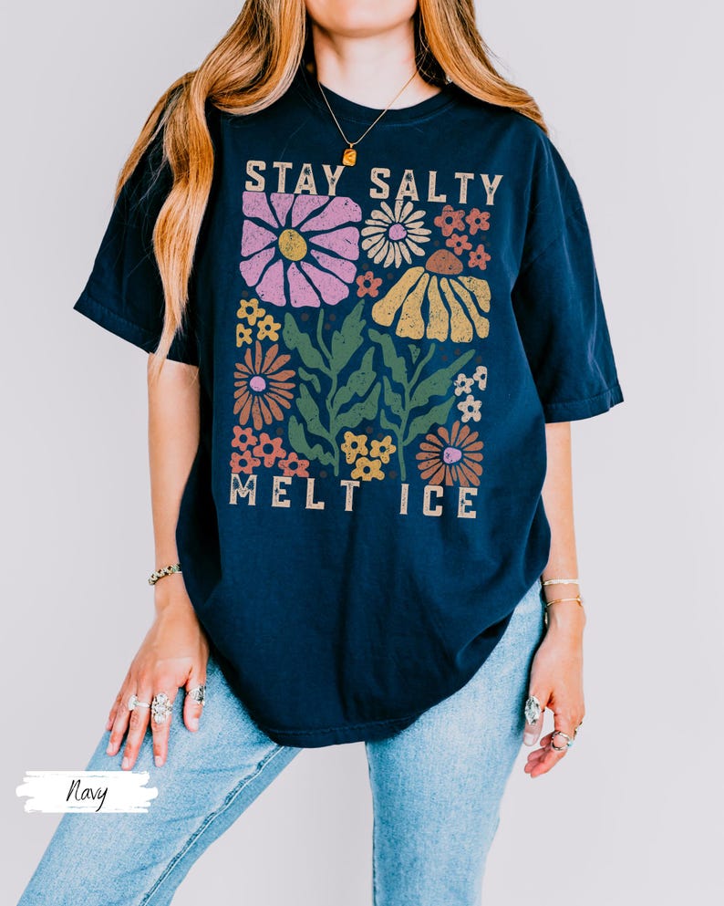 Op de afbeelding: Marineblauwe t-shirt met een bloemenontwerp en de tekst "STAY SALTY MELT ICE". De afbeelding toont roze, gele, oranje en witte bloemen met groene bladeren. Het shirt heeft een relaxte pasvorm.