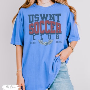 Puede incluir: Una camiseta azul de gran tama&ntilde;o con el texto "USWNT SOCCER CLUB ESTD 1886" en letras rojas y azul marino. La camiseta presenta un gr&aacute;fico de un bal&oacute;n de f&uacute;tbol con alas.