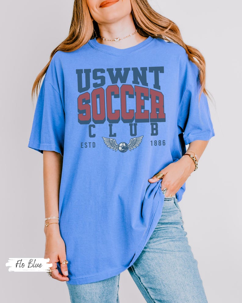 Puede incluir: Una camiseta azul de gran tama&ntilde;o con el texto "USWNT SOCCER CLUB ESTD 1886" en letras rojas y azul marino. La camiseta presenta un gr&aacute;fico de un bal&oacute;n de f&uacute;tbol con alas.