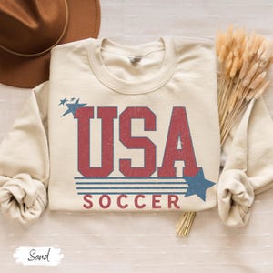 May include: A beige sweatshirt with a vintage-style graphic design that says "USA SOCCER" in red and blue lettering with stars and stripes.