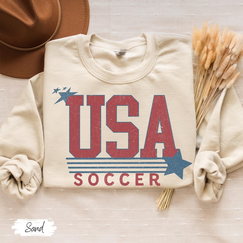 May include: A beige sweatshirt with a vintage-style graphic design that says "USA SOCCER" in red and blue lettering with stars and stripes.