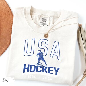 May include: Ivory-colored t-shirt with the text "USA HOCKEY" in blue, featuring a hockey player graphic. The shirt includes the years 1980 and 2026. A gold necklace and a brown hat are visible.