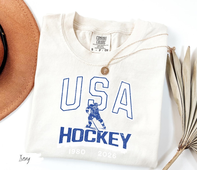 May include: Ivory-colored t-shirt with the text "USA HOCKEY" in blue, featuring a hockey player graphic. The shirt includes the years 1980 and 2026. A gold necklace and a brown hat are visible.