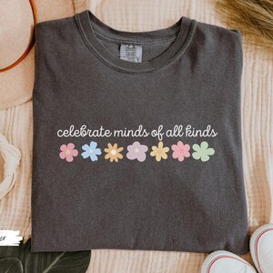 May include: A dark gray t-shirt with the text "celebrate minds of all kinds" printed in white. A row of colorful flowers are printed below the text.
