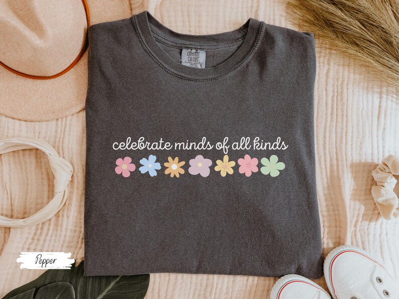 May include: A dark gray t-shirt with the text "celebrate minds of all kinds" printed in white. A row of colorful flowers are printed below the text.