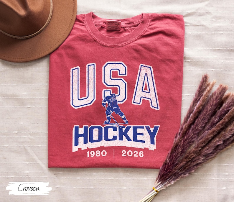 May include: Crimson red t-shirt with the text "USA HOCKEY" in white and blue. A blue hockey player graphic is centered below the text. The years 1980 and 2026 are printed below the word "HOCKEY".
