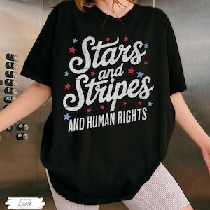 Puede incluir: Camiseta negra con la frase "Stars and Stripes AND HUMAN RIGHTS" en letras blancas, adornada con estrellas rojas y azules. La camiseta es de cuello redondo y manga corta.
