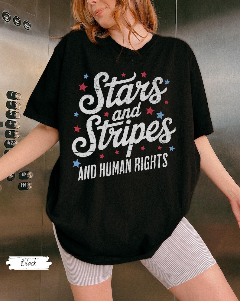 Puede incluir: Camiseta negra con la frase "Stars and Stripes AND HUMAN RIGHTS" en letras blancas, adornada con estrellas rojas y azules. La camiseta es de cuello redondo y manga corta.