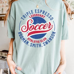 Puede incluir: Una camiseta azul claro con un dise&ntilde;o gr&aacute;fico de estilo vintage. El gr&aacute;fico presenta un c&iacute;rculo azul con el texto "Triple Espresso Legendary Soccer Rodman, Smith, Swanson Athletic Club" en contorno blanco. La palabra "Soccer" est&aacute; en letras rojas y azules.