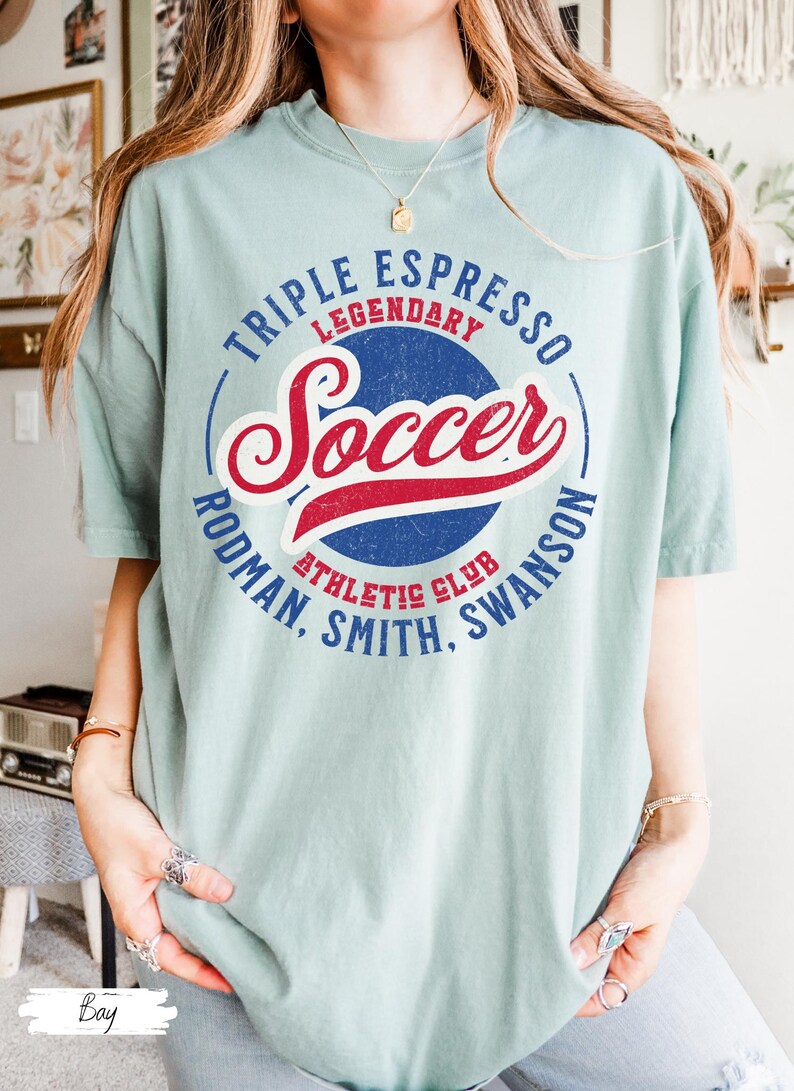 Puede incluir: Una camiseta azul claro con un dise&ntilde;o gr&aacute;fico de estilo vintage. El gr&aacute;fico presenta un c&iacute;rculo azul con el texto "Triple Espresso Legendary Soccer Rodman, Smith, Swanson Athletic Club" en contorno blanco. La palabra "Soccer" est&aacute; en letras rojas y azules.