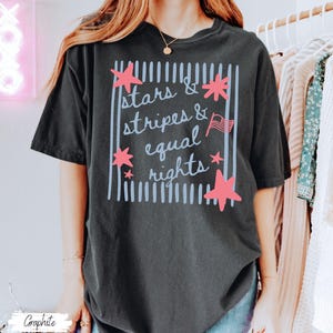 Puede incluir: Camiseta de color grafito con el texto "Stars & Stripes & Equal Rights" en un dise&ntilde;o rectangular. El dise&ntilde;o incluye estrellas rosas y una peque&ntilde;a bandera americana. La camiseta es de estilo c&oacute;modo e informal.