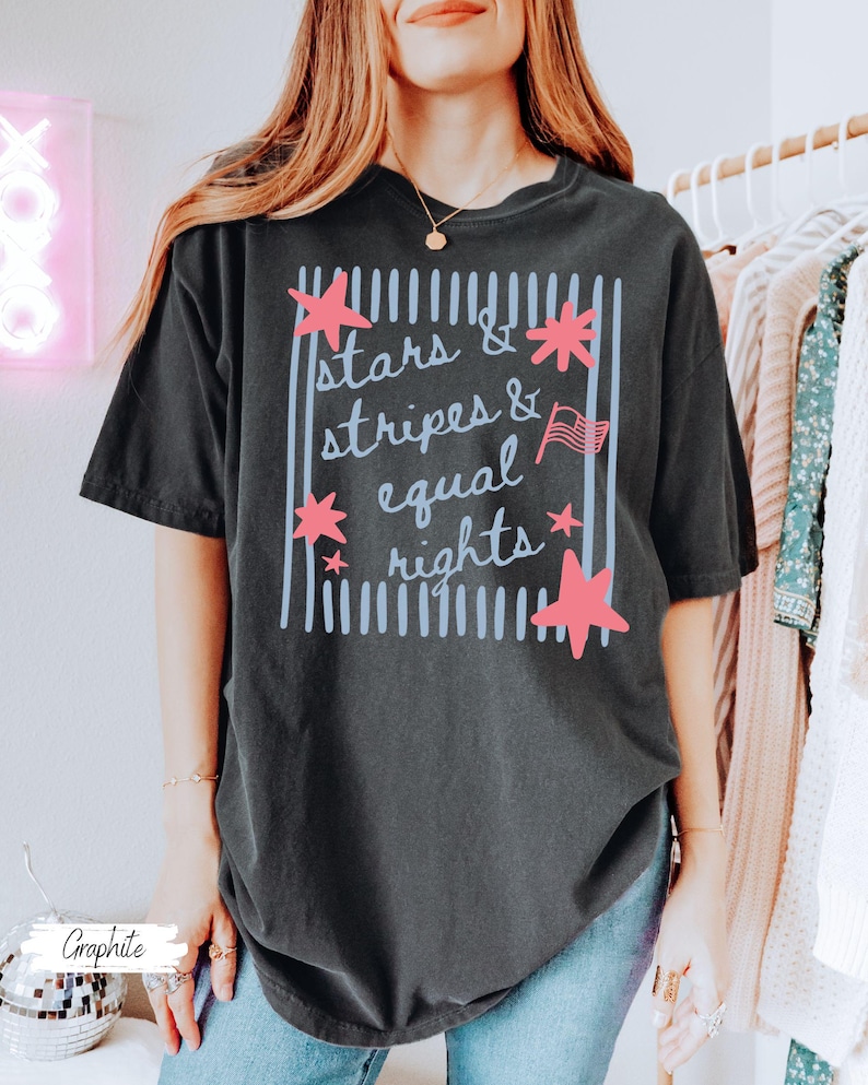 Puede incluir: Camiseta de color grafito con el texto "Stars & Stripes & Equal Rights" en un dise&ntilde;o rectangular. El dise&ntilde;o incluye estrellas rosas y una peque&ntilde;a bandera americana. La camiseta es de estilo c&oacute;modo e informal.