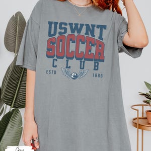 Puede incluir: Una camiseta gris de gran tama&ntilde;o con el texto "USWNT SOCCER CLUB ESTD 1886" impreso en la parte delantera. El texto est&aacute; en una fuente de estilo vintage con un esquema de color rojo y azul. La camiseta presenta un gr&aacute;fico de un bal&oacute;n de f&uacute;tbol con alas.
